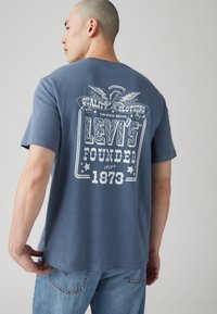 Mėlyna cotton marškinėliai su dideliu baltu grafiniu paveikslu ant nugaros, ant kurio yra užrašas „Levi’s Founded 1873“ ir erelio dizainas; trumpomis rankovėmis ir apvaliu kaklu.