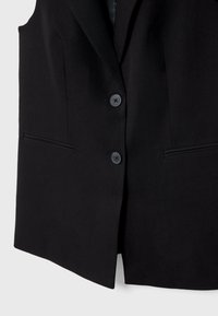 Musta hihaton bleiseri, tehty teksturoidusta kankaasta, jossa on yksinkertainen etusuljenta kahdella napilla ja kaksi sivutaskua.