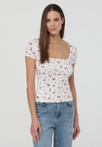 T-shirt bianco in cotone con scollatura quadrata, maniche corte e motivo floreale con rose rosa. Indossato con jeans a vita alta blu.