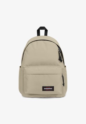 Beige stoffen rugzak met een ronde vorm, zwarte ritsen en een voorvak. Heeft een handvat aan de bovenkant en een logo patch aan de voorkant.