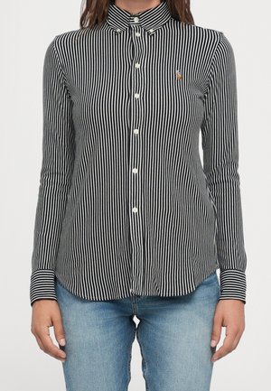 Kvinde iført en tætsiddende sort-hvidt, vertikalt striped knappet skjorte med lange ærmer og lyseblå jeans.