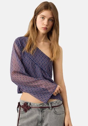Bluza cu un umăr dezgolit, de culoare bleumarin, cu un model geometric texturat, având o mânecă lungă transparentă, asortată cu blugi gri deschis și o curea decorativă.