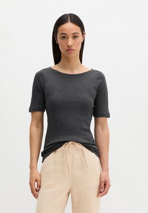 Kobieta o prostych ciemnych włosach, ubrana w dopasowany ciemnoszary krótki rękaw i luźne beżowe spodnie ze ściągaczem, stojąca na jednolitym tle.