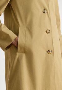 Khaki trenčkot s hladkou textúrou, vybavený bočnými vreckami, výraznými zlatými gombíkmi a priliehavým strihom. Zapnutý a mierne rozopnutý.