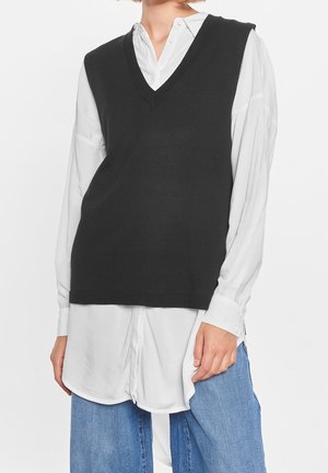 Woman wearing sleeveless black V-neck sweater vest over a long white button-up shirt with blue jeans, standing against white background.