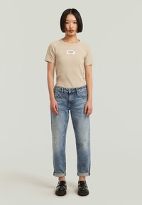 T-shirt en coton beige avec un patch logo carré, associée à un jean bleu clair déchiré avec des ourlets retroussés et des mocassins noirs.
