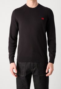 Pull noir en maille avec un col rond, des poignets et une ceinture côtelés, présentant un petit patch logo rouge sur la poitrine. Porté avec un jean foncé.