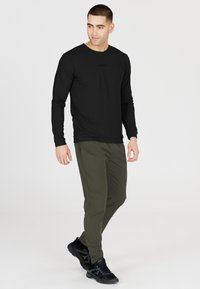 Schwarzes Langarmshirt mit geripptem Textur und kleinem Logo, kombiniert mit olivgrünen, taillierten Hosen und schwarzen Sportschuhen.