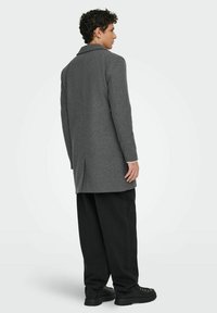 Grahamfarbener Wollmantel mit tailliertem Kragen und langen Ärmeln, ausgestattet mit einem einzelnen Schlitz auf der Rückseite, kombiniert mit lockeren schwarzen Hosen und schwarzen Stiefeln.