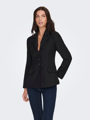 Femme aux longs cheveux foncés portant un blazer noir ajusté à trois boutons et un jean bleu foncé, debout devant un fond clair uni.