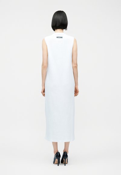 Γυναίκα με κοντά μαύρα μαλλιά που φορά ένα λευκό μίντι φόρεμα χωρίς μανίκια με το κείμενο «Moschino» στο πάνω μέρος της πλάτης και μαύρες ψηλοτάκουνες γόβες.