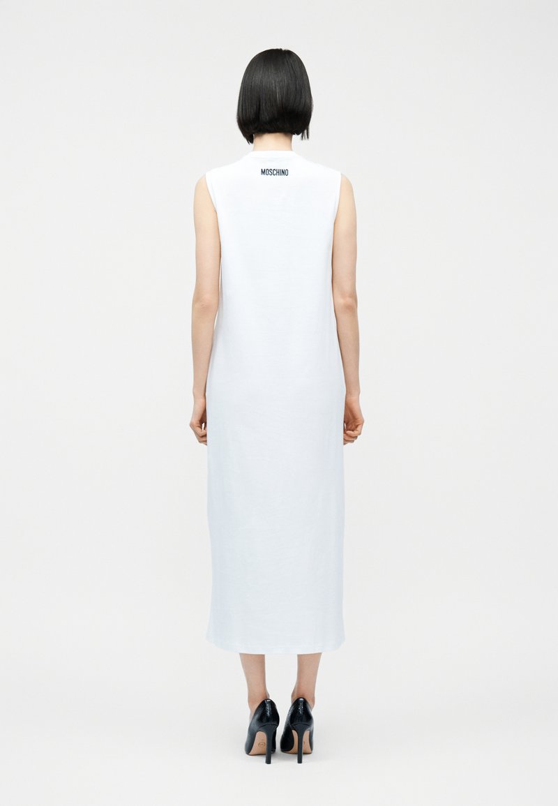 Femme aux cheveux noirs courts portant une robe midi blanche sans manches avec le texte "Moschino" sur le haut du dos et des chaussures à talons noirs.
