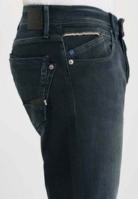 Jean en denim bleu foncé avec une coupe slim, des coutures visibles, plusieurs poches et une étiquette en cuir sur la taille.