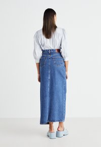 Jupe maxi en denim bleu avec une taille ajustée et deux poches arrière, associée à une chemise bleu clair à manches bouffantes et des mules à talons bleu clair.