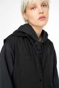 Gilet nero senza maniche con chiusura a bottoni, due tasche sul petto, indossato sopra una felpa con cappuccio scura. Tessuto morbido e opaco senza motivi.