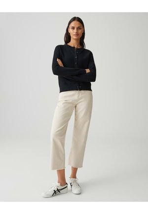 Femme debout, bras croisés, portant un cardigan noir boutonné, un pantalon large crème et des baskets blanches avec des rayures noires.