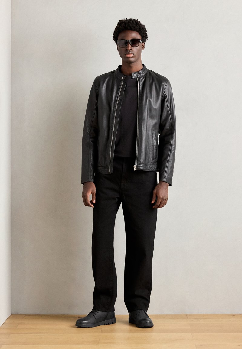 Giacca di pelle nera con una finitura liscia, t-shirt nera, pantaloni neri e scarpe nere. Occhiali da sole e design minimalista.