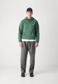 Zelená mikina s kapucí a šňůrkou, přední kapsou a logem; šedé volné kalhoty; černá kšiltovka; černá obuv. Jednoduchý, neformální outfit.