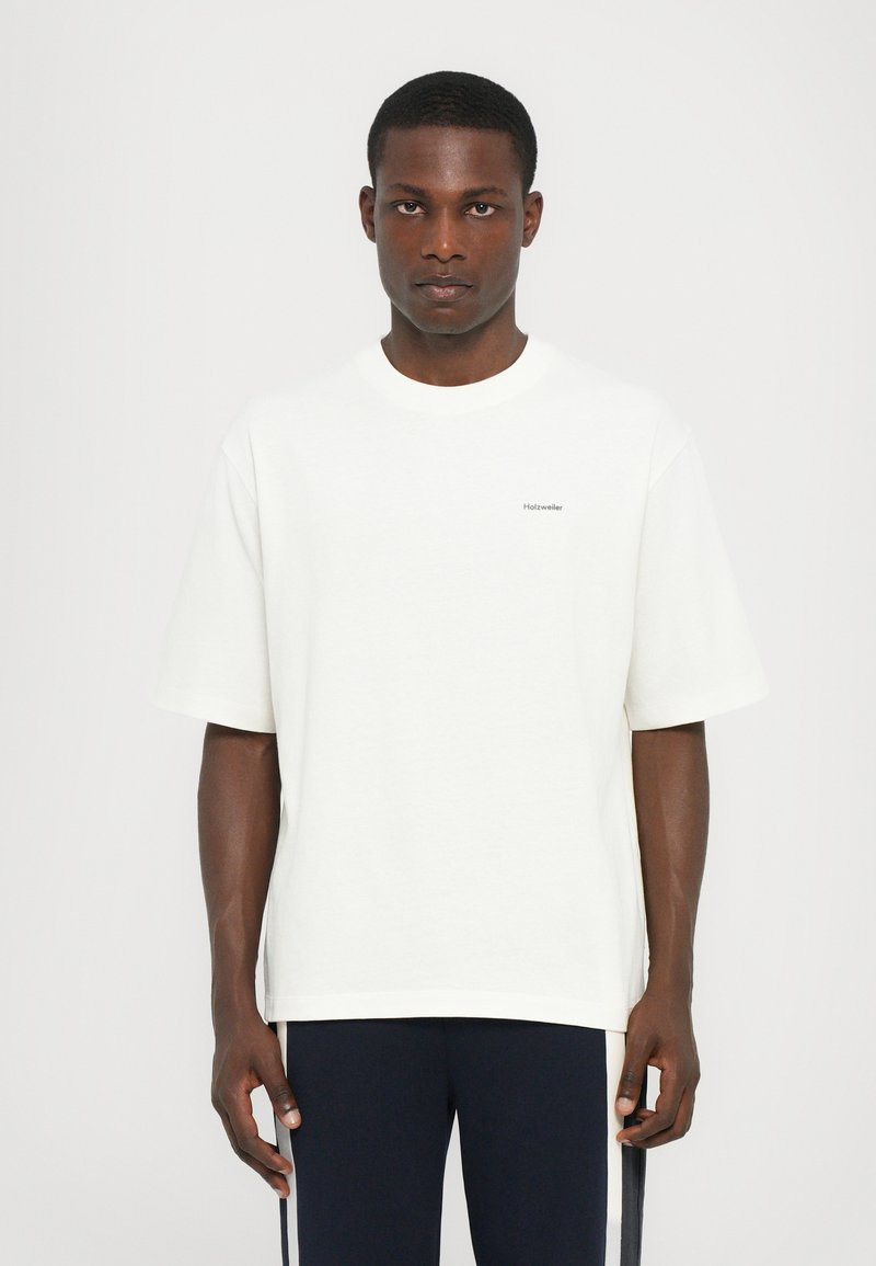 Camiseta de algodón blanca con mangas cortas, corte relajado y un pequeño logo gris en el lado izquierdo del pecho. Textura suave, cuello redondo.