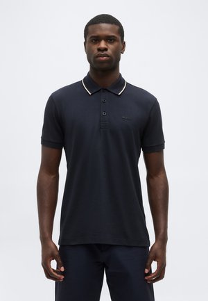 Mann trägt ein dunkelblaues Poloshirt mit kurzen Ärmeln und einem hellen Kontraststreifen am Kragen, steht vor einem einfarbigen Hintergrund.