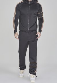 Survêtement de sport noir comprenant un sweat à capuche zippé avec des bandes latérales orange et blanches et un pantalon assorti. Tissu lisse avec poignets élastiques.
