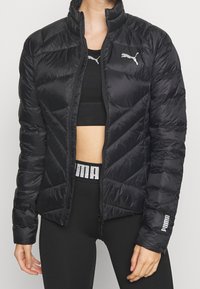 Czarna puchowa kurtka z wysokim kołnierzem, pikowanym wzorem i zapięciem na zamek. Posiada odblaskowe logo na klatce piersiowej i rękawie.
