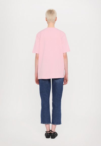 T-shirt oversized rosa, mangas curtas, tecido macio, vista traseira. Combinado com jeans curtos azuis e sapatos pretos com tachas.