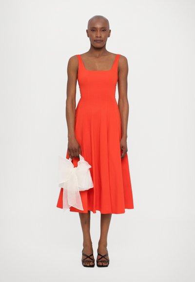 Crvena haljina bez rukava sa kvadratnim izrezom i raširenom suknjom. Drži se bijela prozirna torba s mašnom. Crne sandale s trakama upotpunjuju izgled.