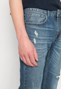 Jeans de denim azul com um acabamento desbotado, detalhes desgastados e ligeiramente desfiados. Apresenta cinco bolsos e uma cintura padrão.