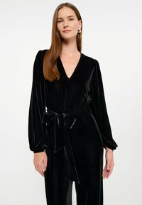 Czarny aksamitny kombinezon z długimi bufkami, dekoltem w kształcie litery V i zwężoną talią podkreśloną pasującym do materiału paskiem. Gładka tekstura.