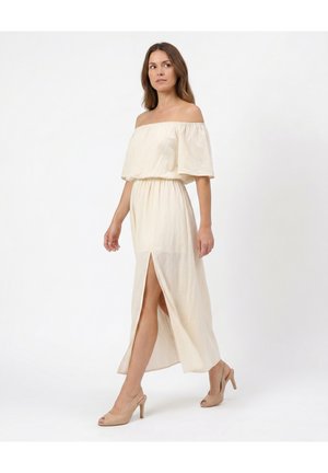 Kvinde iført en cremet off-shoulder kjole med høj slids og beige højhælede sko mod en ensfarvet hvid baggrund.