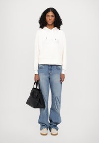 Cappotto corto in crema chiaro con dettagli testuali, jeans baggy blu con lavaggio chiaro e accenti stampati, borsa nera e sneakers color crema.