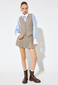 Karierte braune Weste mit Knöpfen, hellblauses Hemd mit übergroßen Manschetten, passender Rock und braune Ankle Boots.