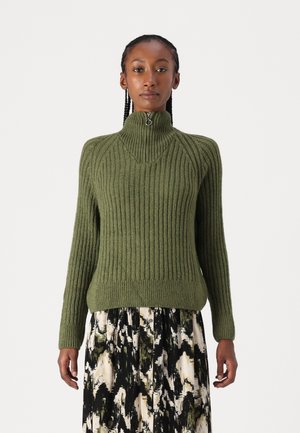 Pull vert à côtes avec un col haut et des détails en zipper, présentant des manches longues et un ourlet ajusté. Associé à une jupe fluide à motifs.
