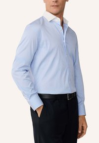 Mann trägt blau-weiß gestreiftes Hemd mit weißem Kragen, schwarzen Gürtel und dunkle Hose, steht mit einer Hand in der Tasche.