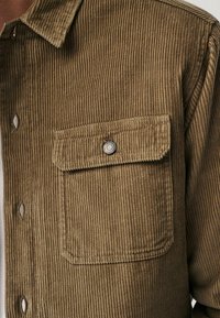 Braunes Cordhemd mit vertikaler Rippstruktur, ausgestattet mit einem Button-down-Kragen und einer Brusttasche mit silbernem Druckknopf.