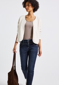 Cardigan en texture crème sur un top beige, associé à un jean bleu foncé. Le mannequin tient un sac en daim marron, mettant en avant une tenue décontractée et superposée.