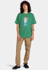 Grøn bomuld t-shirt med et grafisk tryk af en karakter, indrammet af palmer, og ordet "Element" nederst.