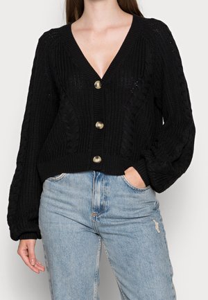 Musta neulekarvattu, jossa on V-pääntie, paksut kabelikuviot ja suuret kultaiset napit. Lyhennetty malli, jossa on rento istuvuus ja leveät hihat.