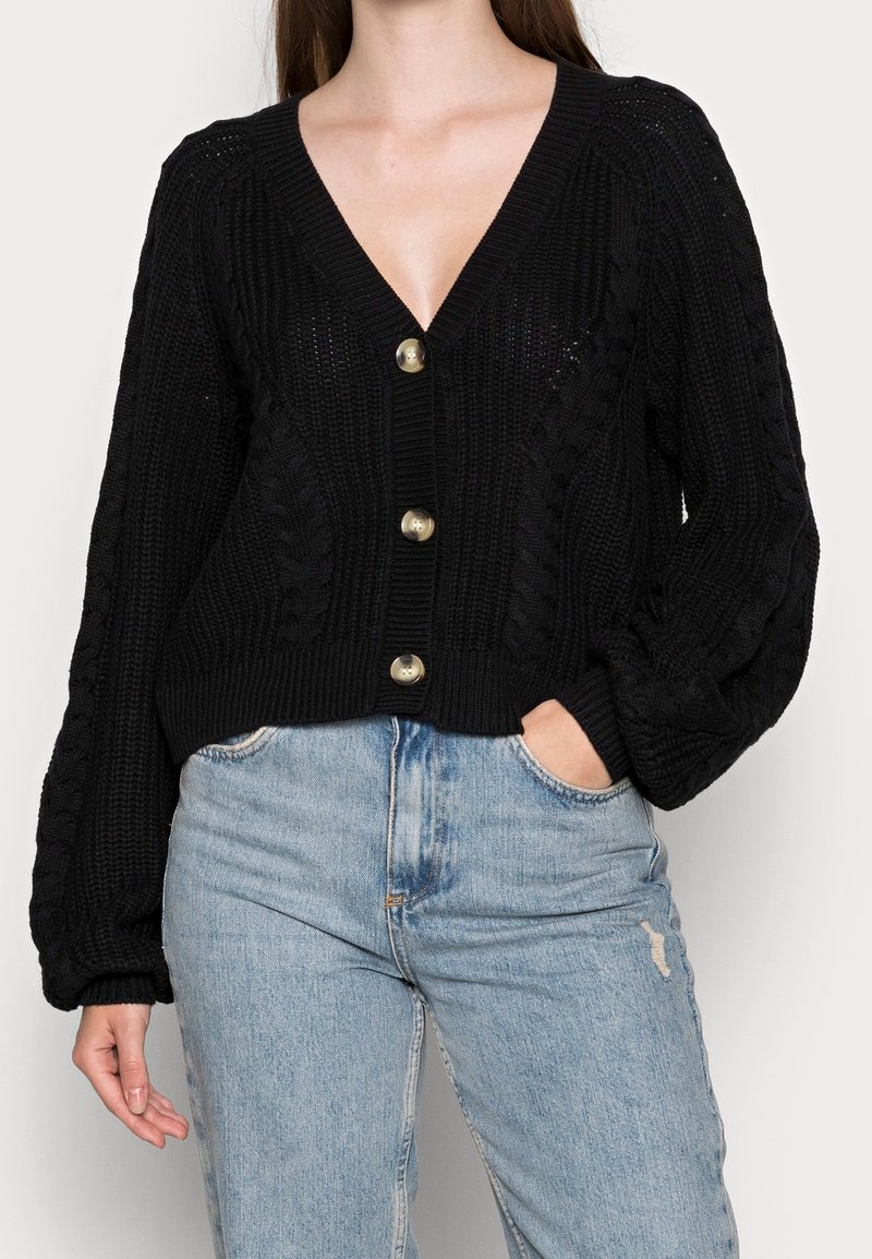 Svart stickad cardigan med V-ringning, prydd med grova kabelmönster och stora guldfärgade knappar. Kort design med en avslappnad passform och vida ärmar.
