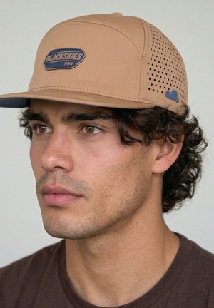 Young man with curly hair wearing a tan Blackskies Pro cap with perforated side panels and a small blue logo.