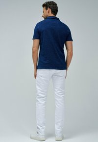 Tummansininen teksturoidulla pinnalla oleva polo-paita, jossa on lyhyt kaulus, yhdistettynä valkoisiin slim fit -housuihin ja valkoisiin lenkkareihin, nähtynä takaa.