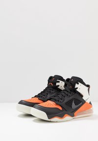 Černé a oranžové tenisky s texturovaným koženým svrškem, síťovinovými detaily a gumovou podešví. Obsahují viditelné polstrování vzduchem a šněrování.