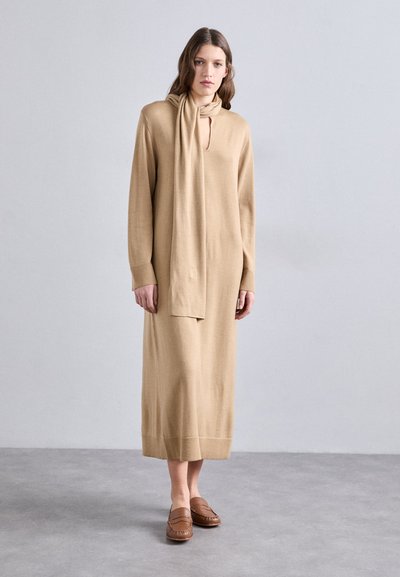 Beige jurk met lange mouwen, een sleutelgat-halslijn en een vastgenaaide sjaal. Zachte textuur, comfortabele pasvorm, reikt tot halverwege de kuit, gecombineerd met een paar bruine schoenen.