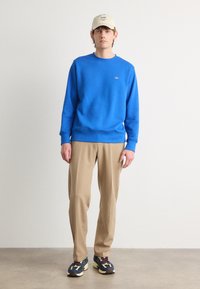 Blauwe sweatshirt, beige broek en meerkleurige sneakers. De sweatshirt heeft een klein logo, een ronde halslijn en geribde manchetten.