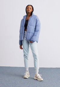 Doudoune bleu clair avec col montant, coupe ample et design matelassé, portée avec un jean en denim clair et des baskets beiges.