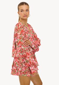 Vrouw met haar in een staart draagt een roze en rode bloemenjurk met lange mouwen, vol met ruches, gouden hoopsieraden en een gouden ketting.