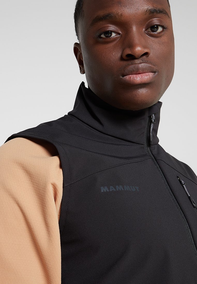 Schwarze Softshell-Weste mit hohem Kragen, Reißverschlusstasche und aufgedrucktem Logo. Über einem beigen Langarmoberteil getragen. Glatte Textur.