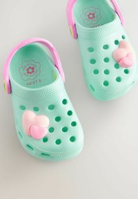 Zuecos moldeados de color verde claro con acentos rosas, decoraciones en forma de corazón, agujeros de ventilación, suelas texturizadas y correas traseras ajustables.