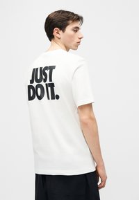 Νέος άνδρας με κοντά καστανά μαλλιά που φοράει λευκό μπλουζάκι με μαύρο κείμενο "JUST DO IT." στην πλάτη, στέκεται μπροστά σε απλό φόντο.